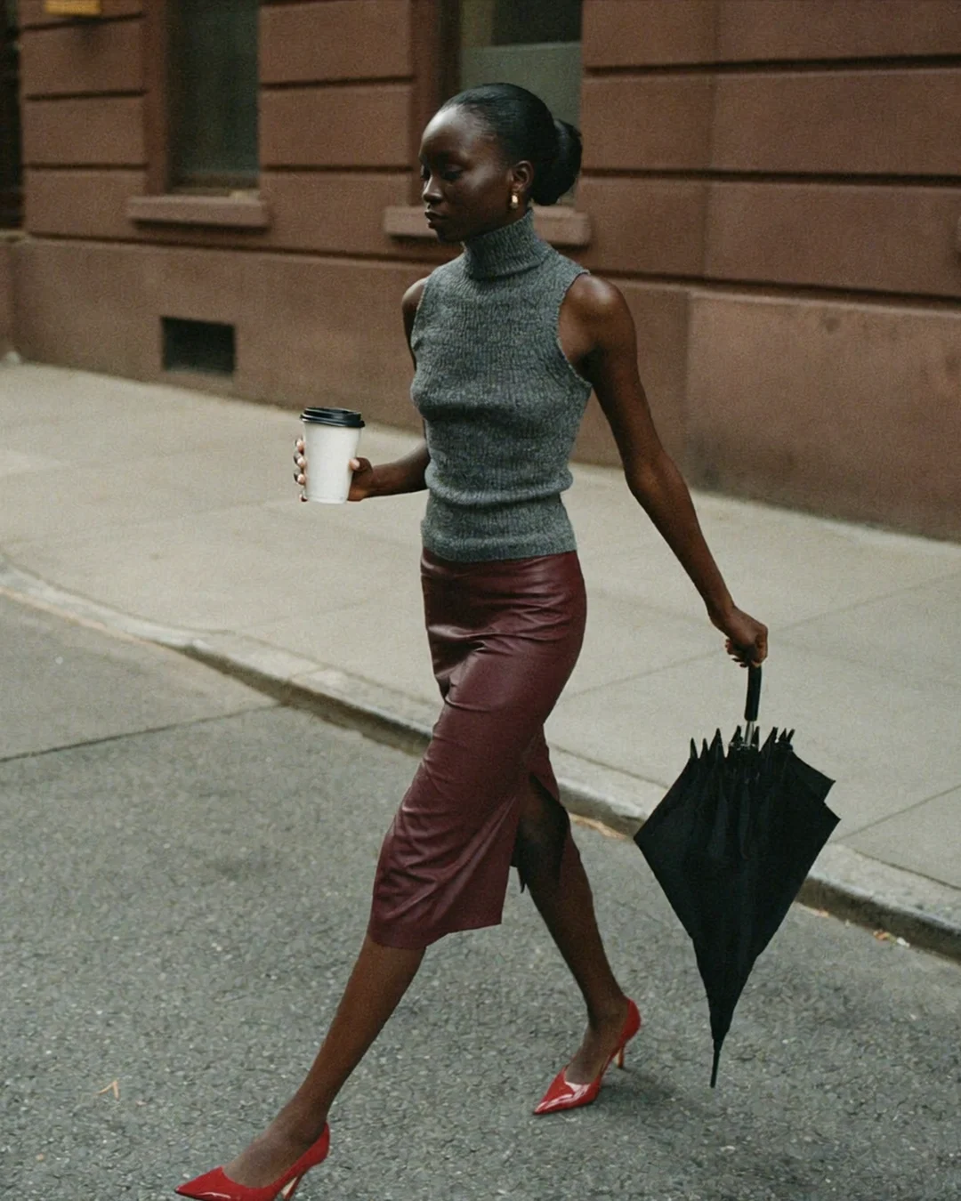 A Black woman in a grey sleeveless turtleneck, burgundy leather pencil skirt and red heels crosses a brownstone street holding a takeaway coffee and a closed black umbrella.