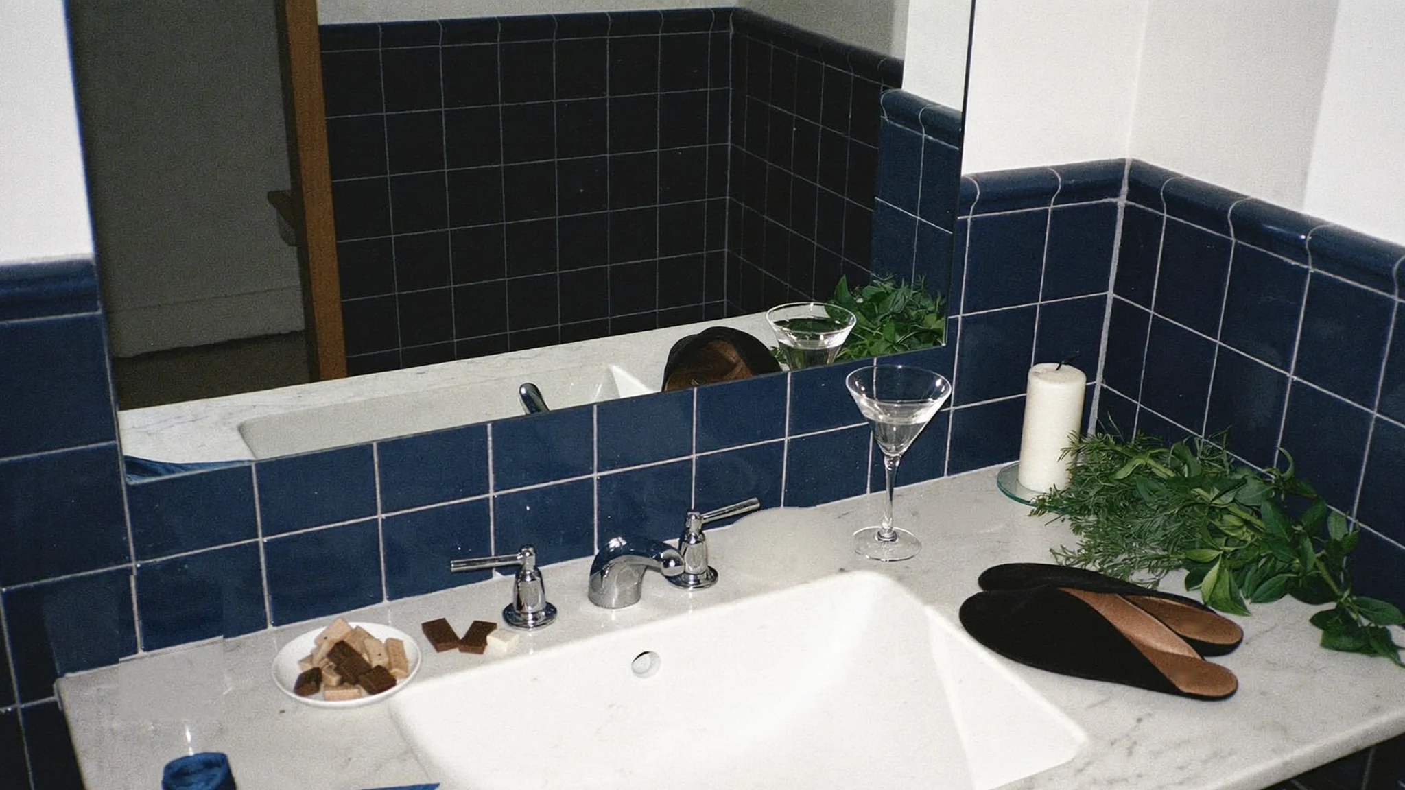 A navy-tiled bathroom vanity with a martini, a white candle, chocolates, black velvet slippers, fresh herbs and a navy silk bow; the Balance blend details are etched into the mirror.