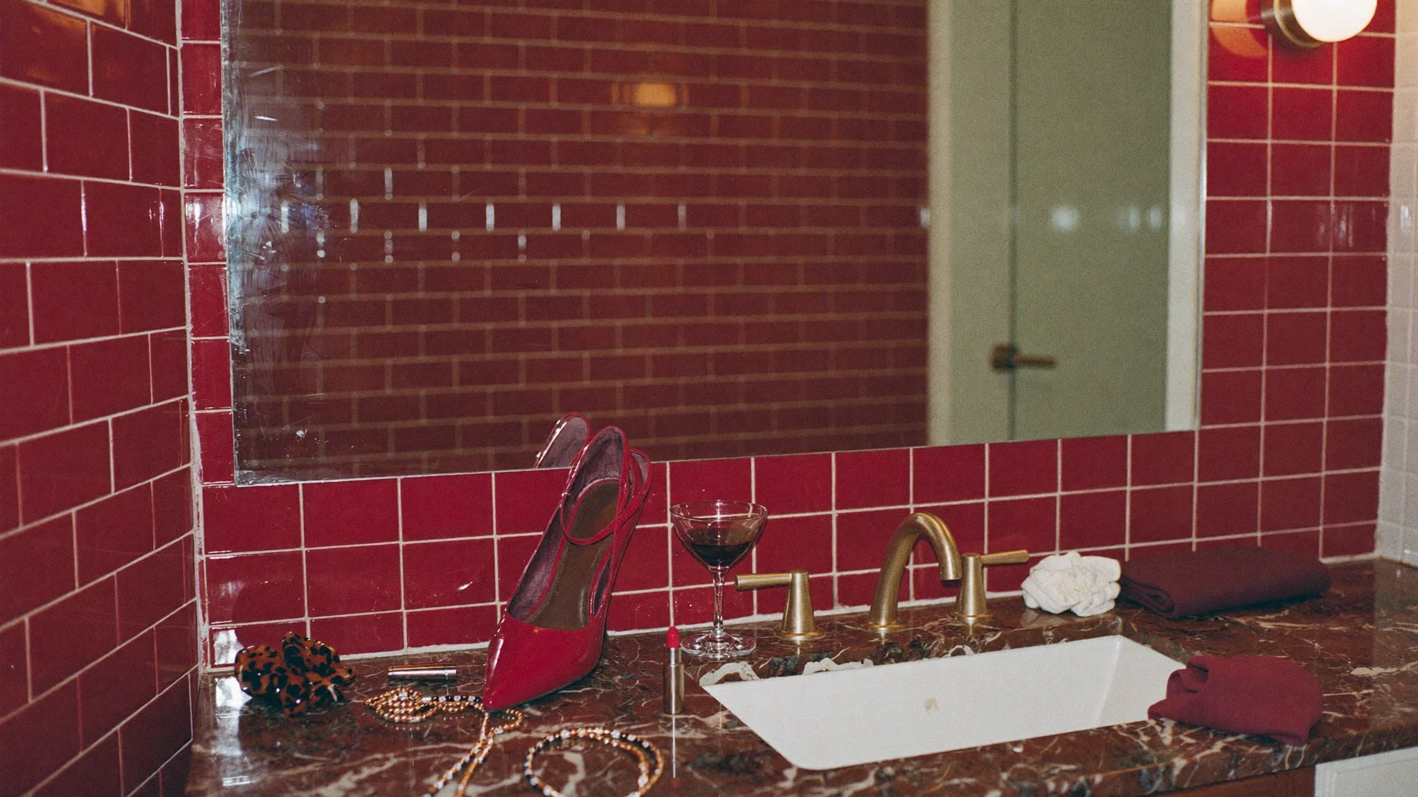 A glossy oxblood-tiled bathroom vanity with red heels, a martini and gold jewelry; the Relief blend details are etched into the mirror.