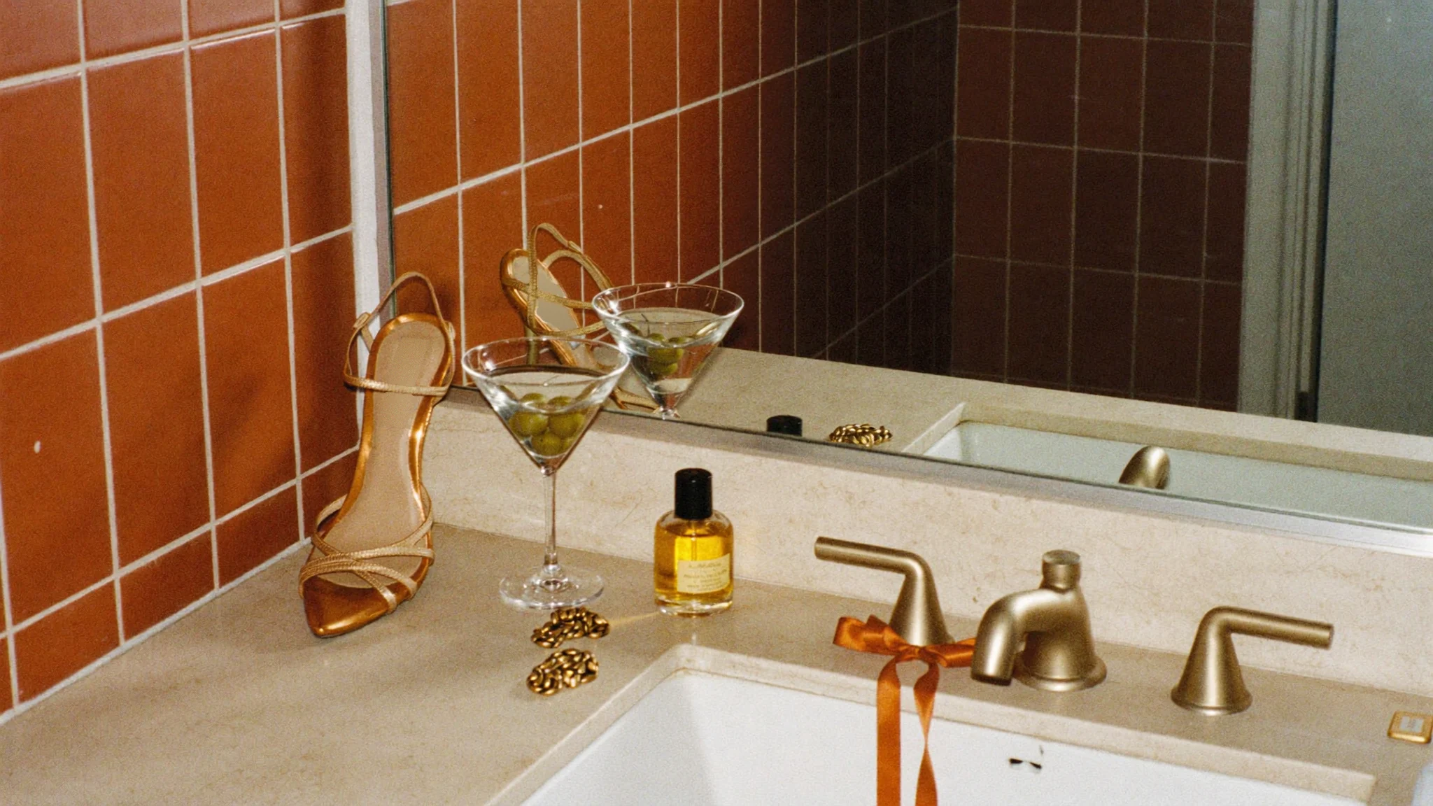 A burnt-orange tiled bathroom vanity with a gold strappy heel, an olive martini, perfume oil, gold chain bracelets and a rust ribbon tied on the gold faucet; the Peak blend details are etched into the mirror.